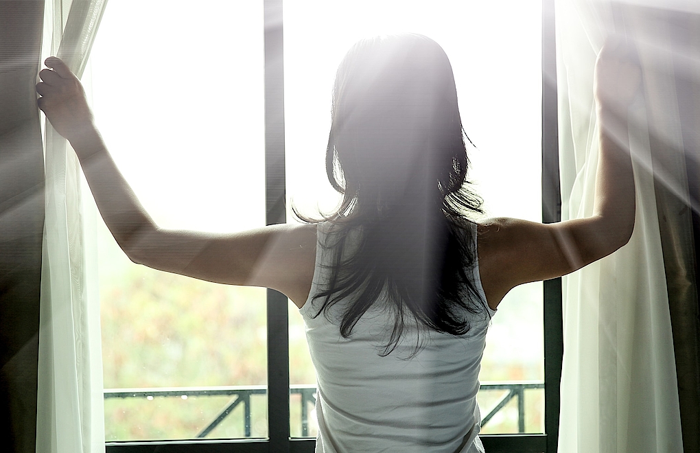 Frau öffnet Vorhänge vor Kunststoff-Alu-Fenster, Sonne strahlt in den Raum