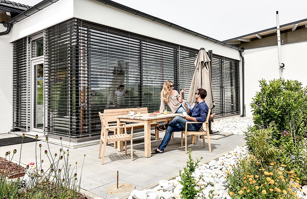 Impulse 100 Strussnig Kunsnstofffenster mit geschlossenem Sonnenschutz von Terrasse aus. Mann und Frau an Terrassentisch