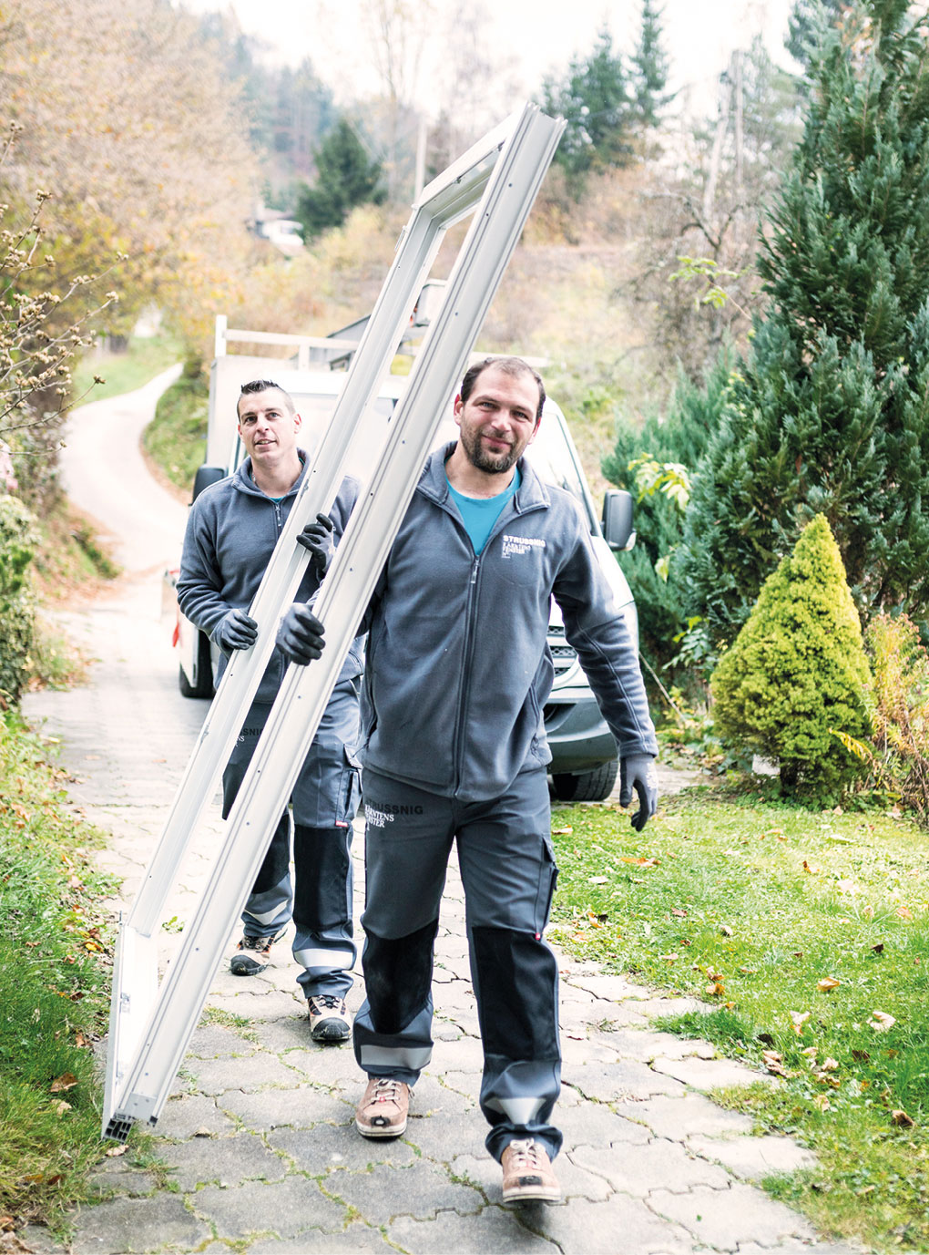 Zwei Herren tragen einen Fensterrahmen, dahinter ist ein Lieferwagen zu sehen.