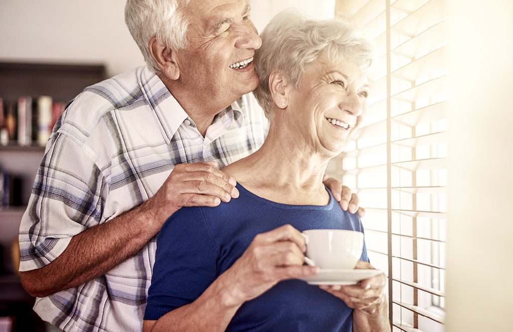 Senior und Seniorin vor lichtdurchflutetem Fenster mit halb geöffneten Faltstores